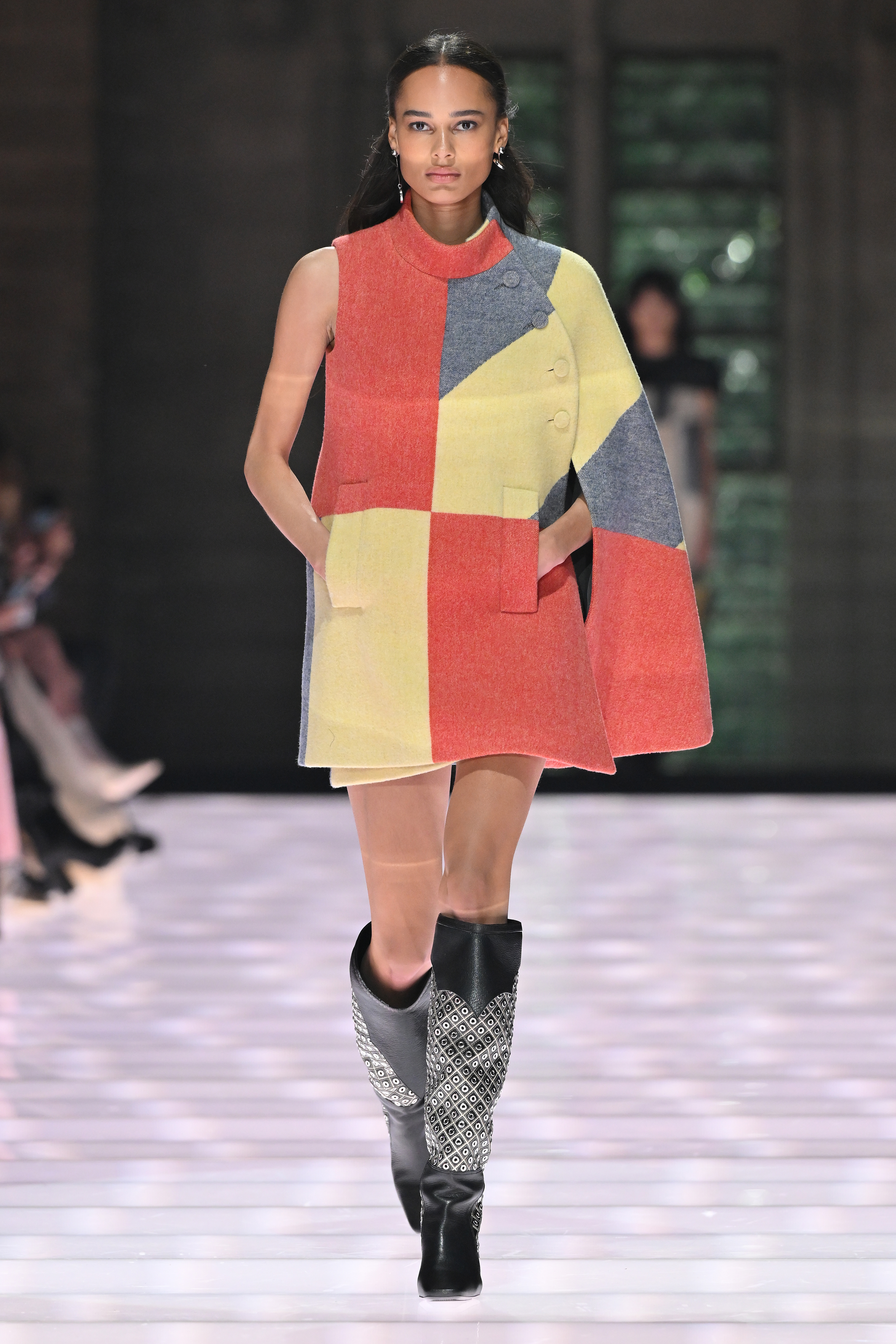 Model walking a runway in a color-blocked cape and tall boots.